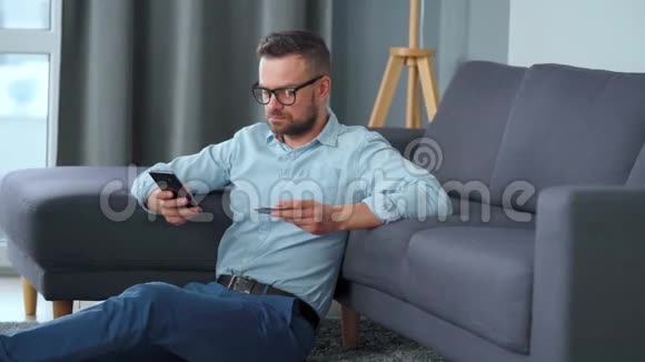 戴眼镜的大胡子坐在沙发附近的地板上用信用卡和智能手机在线付款视频的预览图