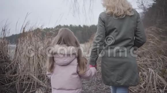 后景白种人妇女和女孩步行到湖边或河流通过灌木丛金发母亲和黑发女儿视频的预览图