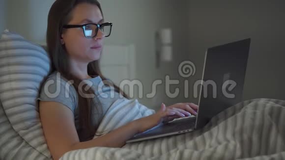 戴眼镜的女人晚上躺在床上用笔记本电脑工作移动成瘾或失眠的概念视频的预览图