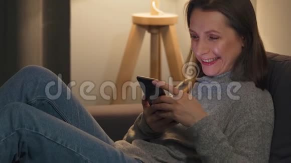 微笑的女人躺在舒适的房间里的沙发上晚上用智能手机上网她非常视频的预览图