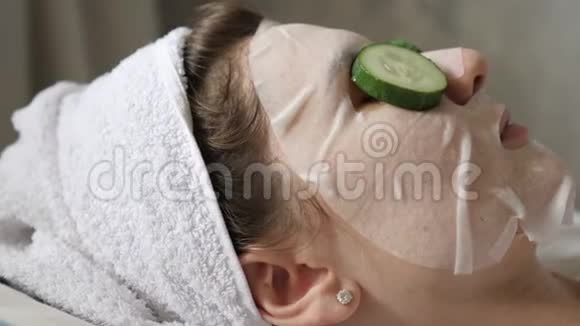 用新鲜黄瓜和保湿面膜进行面部美容治疗头上戴着白色毛巾的女人视频的预览图