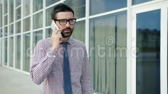 年轻的商人走在办公楼外微笑着用手机说话视频的预览图