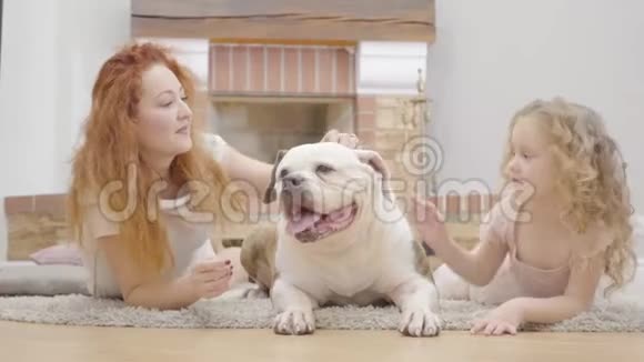 成年红发白种人妇女和小女孩躺在地毯上与宠物牛头犬在家里孩子站起来视频的预览图