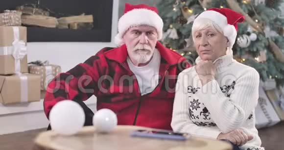 年长夫妇坐在圣诞树旁边看智能手机的肖像等待来自视频的预览图