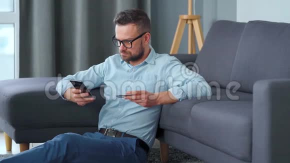 戴眼镜的大胡子坐在沙发附近的地毯上用信用卡和智能手机在线付款视频的预览图