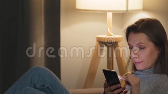微笑的女人躺在舒适的房间里的沙发上晚上用智能手机上网放松和视频的预览图