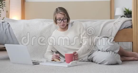 穿着睡衣的女人一边喝咖啡一边吃饼干一边在地毯上用笔记本电脑视频的预览图