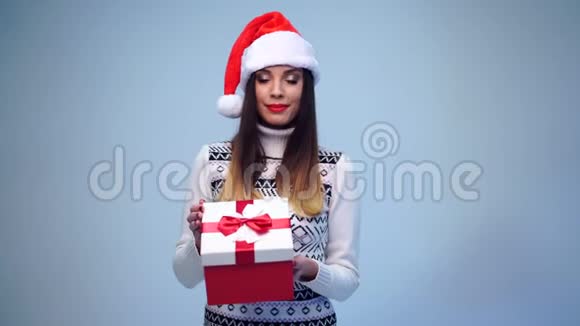 年轻漂亮性感的圣诞老人穿着白色毛衣戴着可爱的圣诞帽打开一个红色的大礼盒视频的预览图