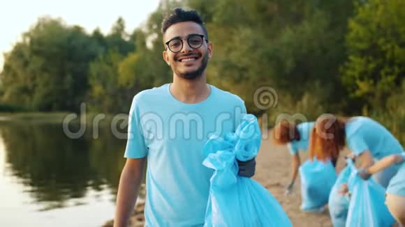 年轻的阿拉伯志愿者带着垃圾袋微笑在湖岸与人的背景视频的预览图