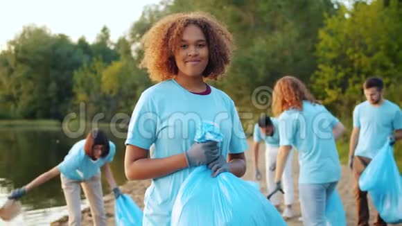 微笑的非裔美国人志愿者拿着垃圾桶包在污染的湖岸视频的预览图