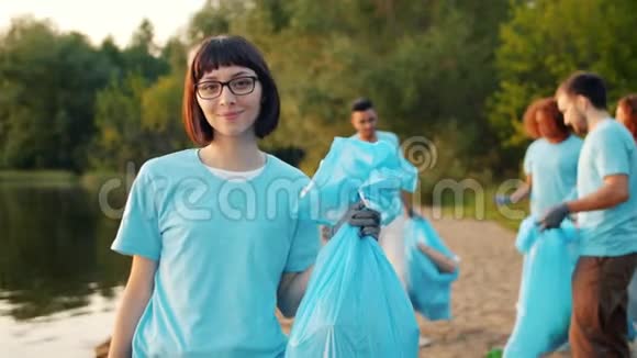 欢乐的女性生态活动家拿着垃圾袋在湖岸与辛勤的团队视频的预览图