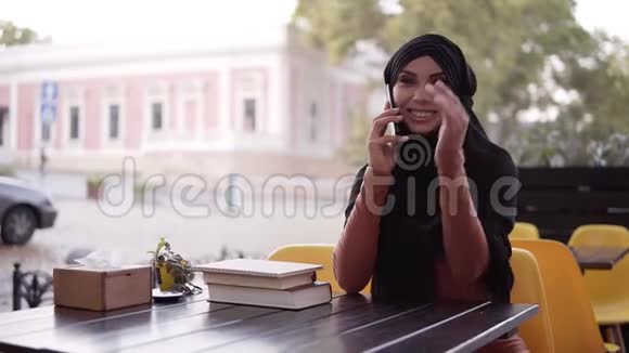年轻漂亮的穆斯林女人戴着头巾在电话里说话微笑着坐在咖啡馆里兴高采烈桌子上有书视频的预览图