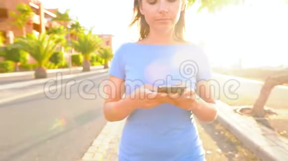 日落时分一个穿着智能手机的蓝色裙子的女人在棕榈街散步视频的预览图