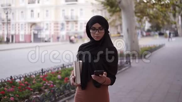 一位年轻的穆斯林妇女戴着传统的头巾背着她街上的智能手机上和朋友聊天视频的预览图