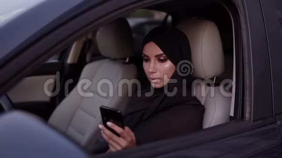 漂亮的年轻女人戴着黑色头巾坐在她的车里沉思地看着她的智能手机屏幕聊天或视频的预览图