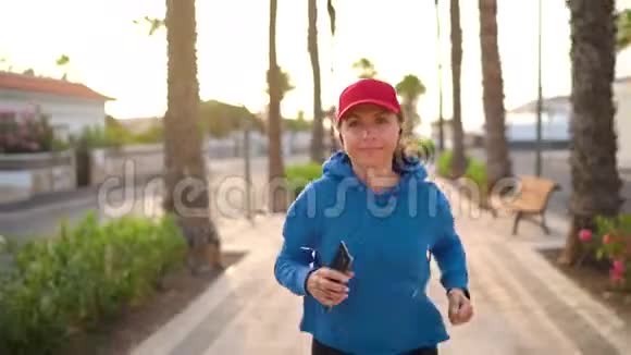 日落时分带着耳机和智能手机的女人沿着棕榈大道沿着街道跑健康的生活方式视频的预览图