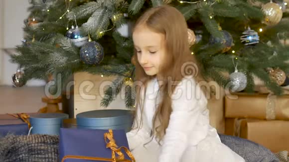 可爱的金发女孩收到了一个礼品盒包裹着工艺深蓝色的纸坐在圣诞树附近童年快乐视频的预览图