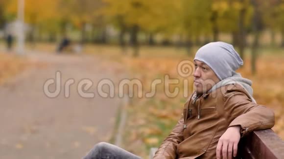 戴着灰色帽子的帅哥正坐在秋天公园的长凳上休息看着人们在公园里散步视频的预览图