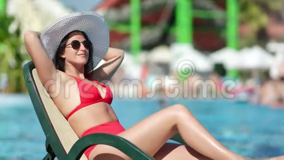 游泳池边的躺椅上戴着帽子穿着红色泳衣的微笑放松的女性视频的预览图