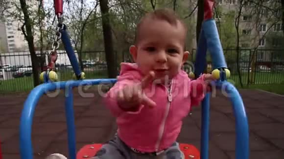 一个小婴儿在秋千上摆动试图用手指伸手去拿相机穿着粉色毛衣和白色衣服的女孩视频的预览图
