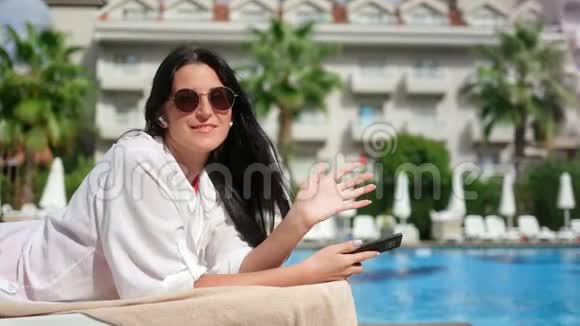 在蓝水棕榈酒店背景下一位快乐的旅行女性在甲板椅上摆着摆手的肖像视频的预览图