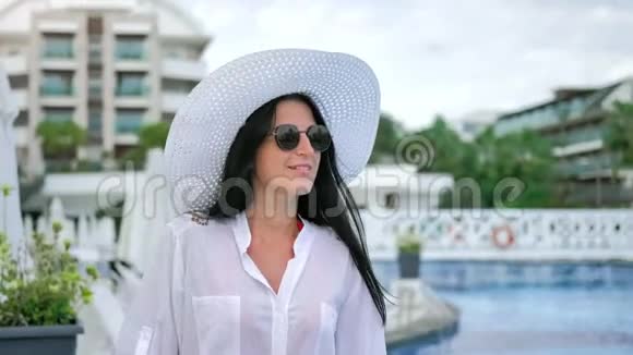 戴着帽子戴着墨镜的微笑旅游妇女在游泳池旁漫步欣赏豪华度假胜地视频的预览图