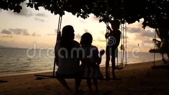 度假的幸福家庭爸爸妈妈和宝宝骑在秋千上看海边的日落视频的预览图