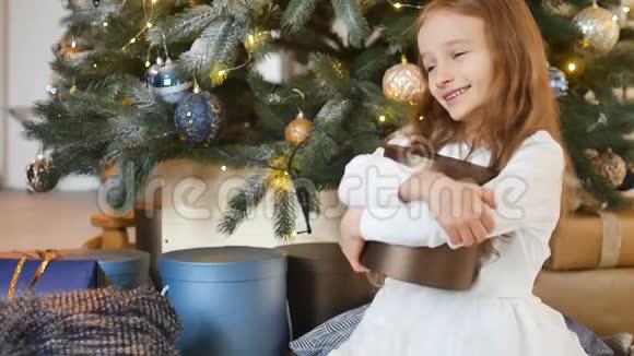 微笑着可爱的金发女孩拥抱着一个金色的礼盒她坐在圣诞树旁快乐的童年和圣诞节视频的预览图