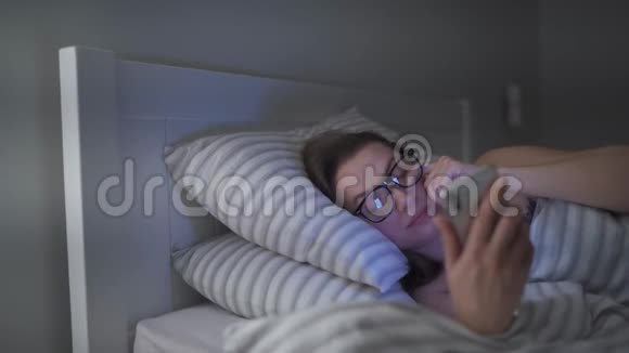 戴眼镜的女人躺在床上使用智能手机她揉眼睛因为她又累又困移动视频的预览图
