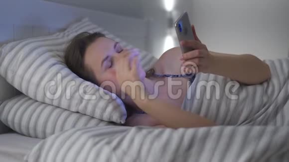 女人躺在床上使用智能手机她揉眼睛因为她又累又困移动成瘾者或视频的预览图