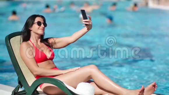 性感的女游客穿着红色泳衣躺在躺椅上在电话上摆出笑的姿势视频的预览图