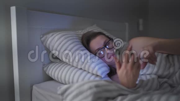 戴眼镜的女人躺在床上使用智能手机她在这个过程中睡着了因为她很累视频的预览图