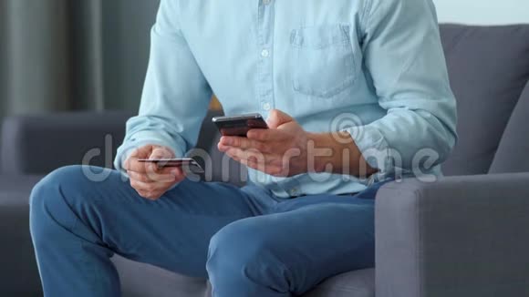 正式穿着的男人坐在沙发上把信用卡号码输入智能手机在线支付网上购物视频的预览图