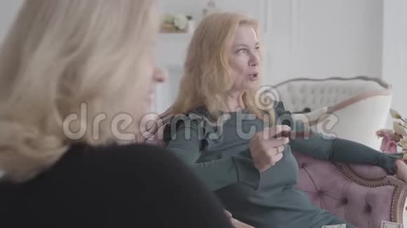 微笑着成熟的高加索女人坐在舒适的扶手椅上拿着钱和雪茄说话老年女性朋友视频的预览图