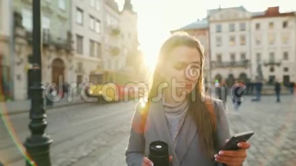 女人手里拿着一个保温杯在夕阳下用智能手机沿着一条老街走着通信社交网络视频的预览图