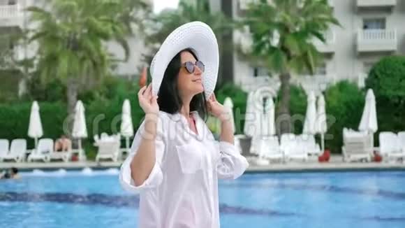 漂亮迷人的女性戴着白帽子戴着太阳镜漫步豪华酒店露台享受假期视频的预览图