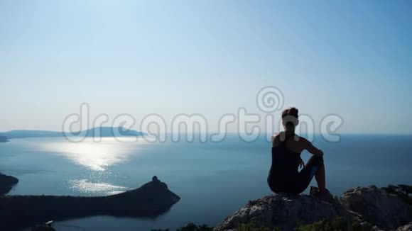 一个快乐的徒步旅行者女人坐在山顶上放松的剪影在克里米亚海湾欣赏美妙景色的女孩视频的预览图