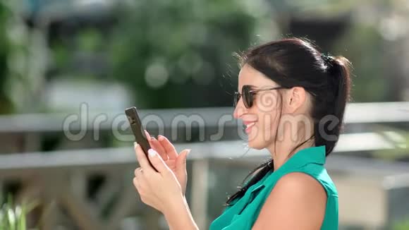 微笑可爱的黑发女孩使用智能手机在夏季公园玩有趣的中型特写视频的预览图