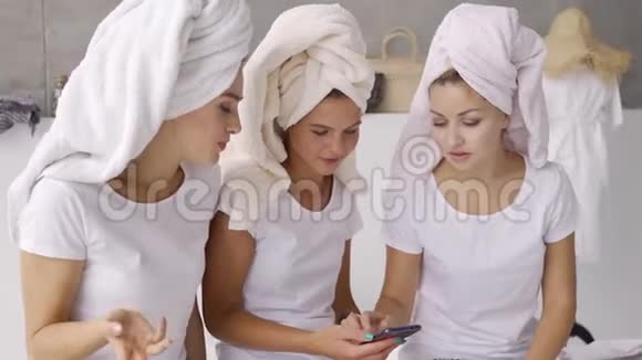 三个积极的女朋友穿着白色T恤衫头上有毛巾一起在家里滚动屏幕视频的预览图