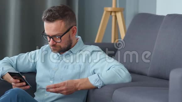戴眼镜的大胡子坐在沙发附近的地板上用信用卡和智能手机在线付款视频的预览图