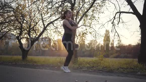 一个年轻的白种人女孩在秋天公园的跳绳锻炼日落户外黑人运动服训练的女子视频的预览图