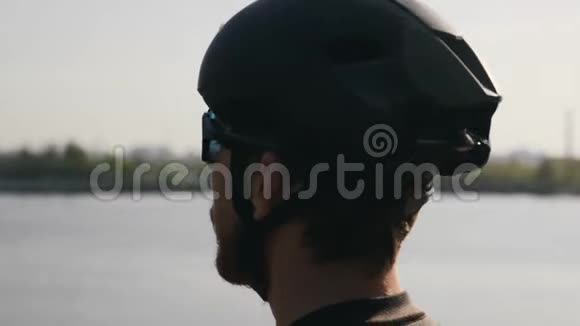 戴着黑色头盔和太阳镜的有胡子的骑自行车的人目光集中到地平线上河和桥梁的背景骑自行车视频的预览图