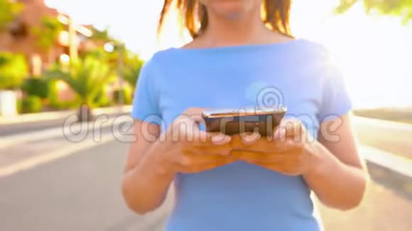 日落时分一个穿着智能手机的蓝色裙子的女人在棕榈街散步视频的预览图
