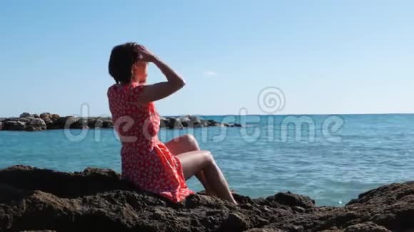年轻苗条的白种人女孩穿着红色短裙坐在大海上的大石头上有魅力的黑发女人直发坐在r附近视频的预览图