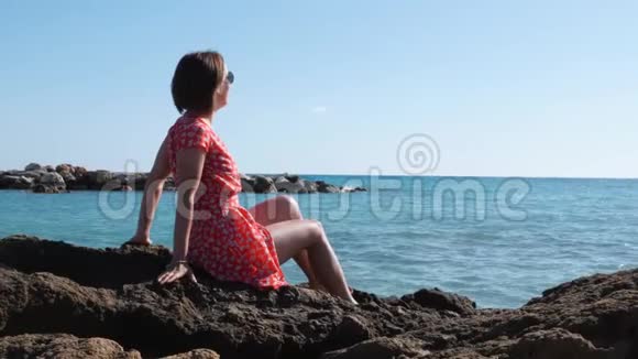美丽的黑发白种人女孩坐在悬崖边被海包围穿红裙子戴太阳镜的年轻女孩坐在视频的预览图