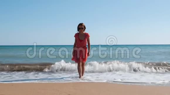 穿着红色连衣裙和黑色太阳镜在沙滩上行走的白人黑发女孩迷人的女人从海上走到摄像机前女孩c视频的预览图