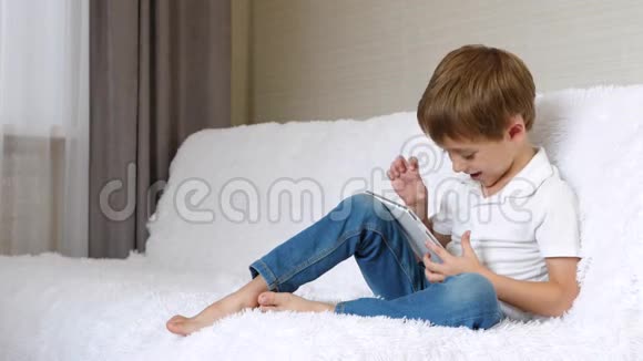 男孩坐在床上用平板电脑在网上玩或看电影孩子感受到了视频的预览图
