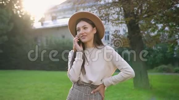 微笑着戴着米色帽子的漂亮年轻女子站在房子院子里的绿树旁打着电话视频的预览图