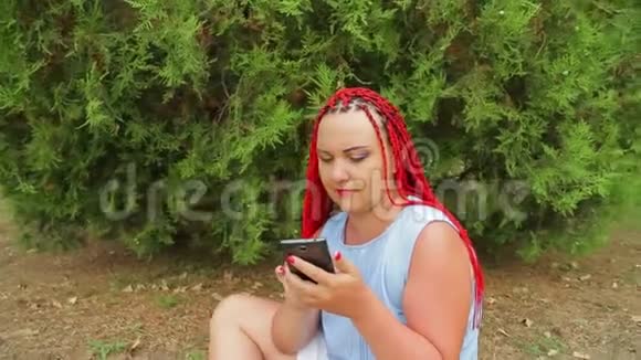 一个年轻的女人坐在草地上的公园里看着她的手机移动摄像机视频的预览图