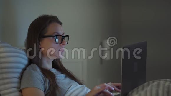 戴眼镜的女人晚上躺在床上用笔记本电脑工作移动成瘾或失眠的概念视频的预览图